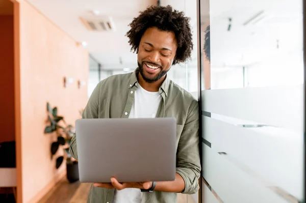 Curly young african-american businessman standing in office hallway and using laptop for office work, male employee checks mail, conducts business correspondence with customers
