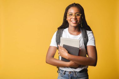 Nice attractive young 20s student girl in basic white t-shirt and glasses standing with backpack and carrying books, bookworm, wiseacre isolated on yellow,