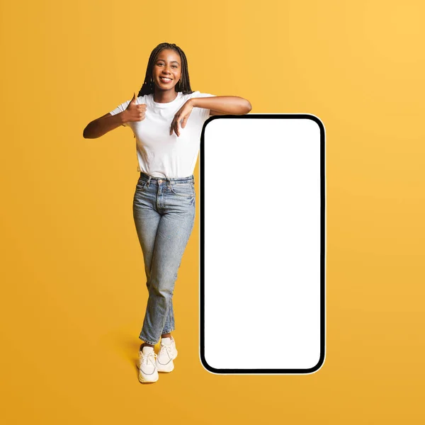 Best deal, promotion. Smiling positive african-american lady leaning on huge smartphone with empty blank screen, showing thumb up, female student recommending new app or website, mockup, isolated