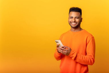 Photo of handsome cheerful smiling man holding mobile phone in the hand and smiling, isolated over yellow background, chatting using smartphone in front camera