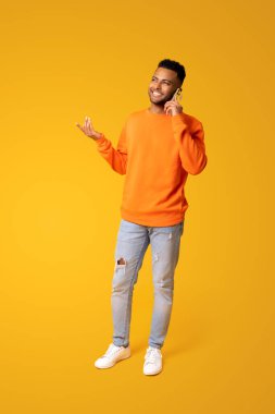 Gesturing indian man talking phone, calling to somebody, looking away with happy smile. Indoor studio shot isolated on yellow background