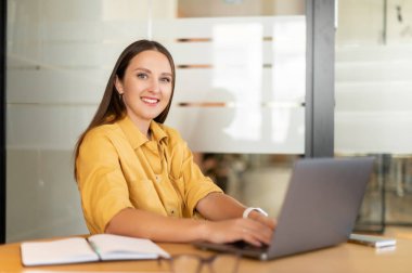 Positive female office worker sitting at the desk, working on project, typing on the laptop, looking at camera with happy smile. Business concept