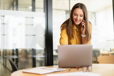 Bossy businesswoman has phone conversation working with laptop at the desk in office. Saleswoman talking with a customers by smartphone. Smiling woman speaking on smartphone