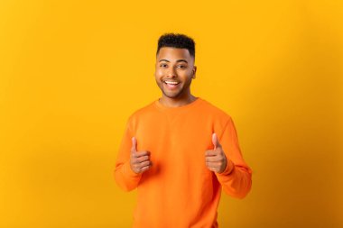 Portrait of man wearing orange casual style sweater, pointing finger pistols to camera playfully and flirting, choosing lucky winner. Indoor studio shot isolated on orange background