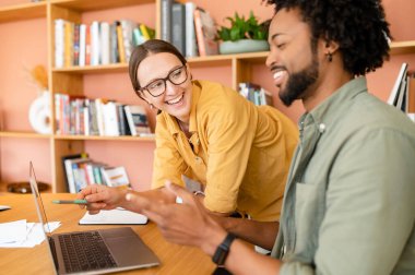 Pair of friendly colleagues work in office. Young businessman explains to his female colleague something using a laptop, they are looking on the screen
