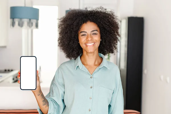 Happy young woman with afro hairstyle showing smartphone with empty screen with apartment interior on background, cheerful housewife recommending mobile app for shopping or quik delivering to the door