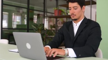 Excited lucky hispanic young businessman sitting at the desk in front of laptop in office and rejoicing with good news, overjoyed male office employee celebrating job offer, clenched fists in triumph