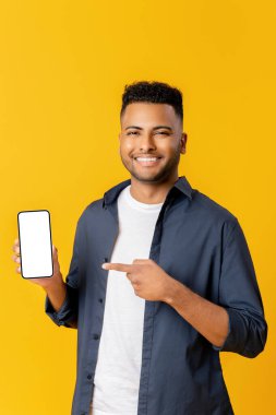 Positive indian man with pointing finger at smartphone with white empty display, bragging with application. Indoor studio shot isolated on yellow background