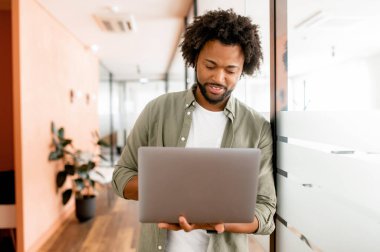 Serious concentrated determined young bearded office worker in casual clothes, stands in office hallway, typing on laptop, freelancer is working on project, writing email to client