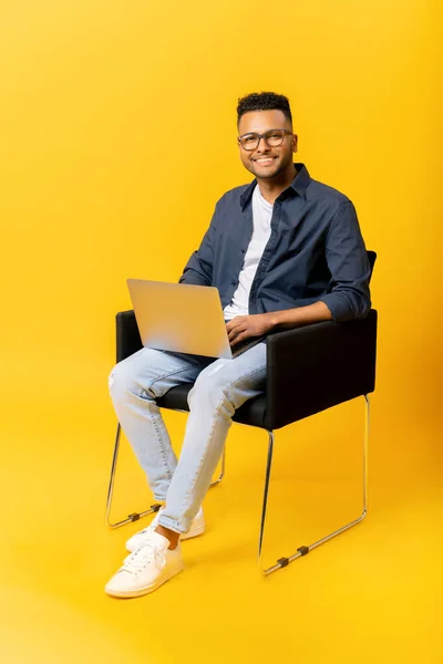 Cheerful indian guy using laptop in casual shirt and glasses using laptop sitting on the chair isolated on yellow, freelancer guy is working remotely, developer coding, student studying, full length