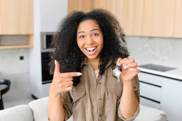 Relocation concept. Headshot of excited female moving in new home or apartment. Cheerful multiracial woman homeowner looking at camera and points finger at the keys with keychain in her hand
