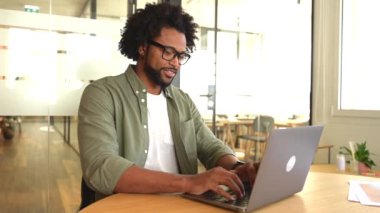 Focused curly handsome man using laptop computer in contemporary coworking space, freelance programmer in glasses coding sitting at the office desk, creativity designer or architect typing
