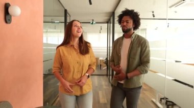 Young enthusiastic female colleague explains new strategy to male employee and gesturing. Indoor portrait of diverse work team discussing, walking through hallway in modern coworking office space