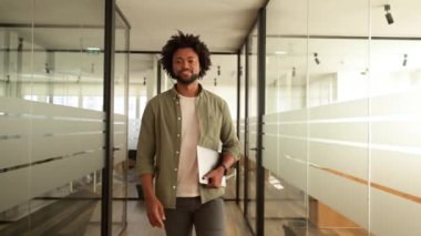 Dertsiz tasasız, modern ofis alanında yürüyen ve elinde dizüstü bilgisayarla gezen Afro-Amerikan erkek çalışanlardan esinlenerek yeşil gömlekli, gururlu iş adamı hafif bir gülümsemeyle kameraya bakar.
