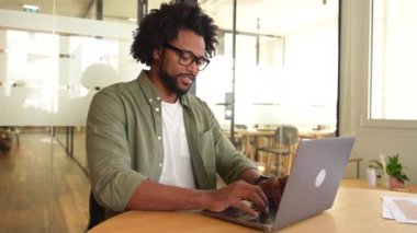 Cheerful african american man wearing casual wear sitting on the workplace and using laptop at modern office. Confident male entrepreneur typing on keyboard