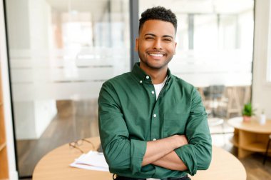 Accomplished Indian male entrepreneur confidently stands with his arms folded, gazing at the camera with a smile. Optimistic and cheerful freelancer in a casual shirt working in shared workspace