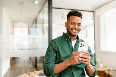 Young Indian male employee, freelancer, businessman stands with a smartphone in hands, beaming with a smile as he looks at the screen, texting and messaging online