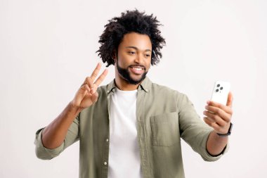 Yakışıklı, çekici 30 'lu yaşlarda, akıllı telefonla selfie çeken beyaz, karizmatik Afrikalı bir adamın görüntülü konuşma yaptığı web kamerasına barış sembolü yerleştiren ve sanal bağlantı için mobil uygulama kullanan bir adam.