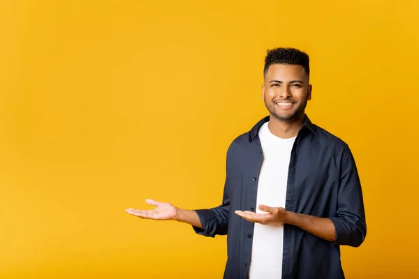 Happy excited young indian man looking at camera pointing aside with both hands, gesturing at copy space advertising new promotion, presenting sale offer standing isolated on yellow background.