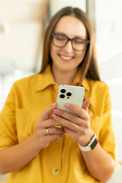 Vertical view of the laughing business lady holding smartphone and looking at the screen while typing message at the office hall. Business people concept