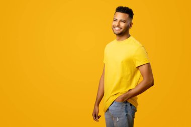 Sarı tişörtlü yakışıklı Hintli genç adam sarı arka planda poz veriyor, dişlek tebessümlü karizmatik adam ayakta duruyor ve yana bakıyor.