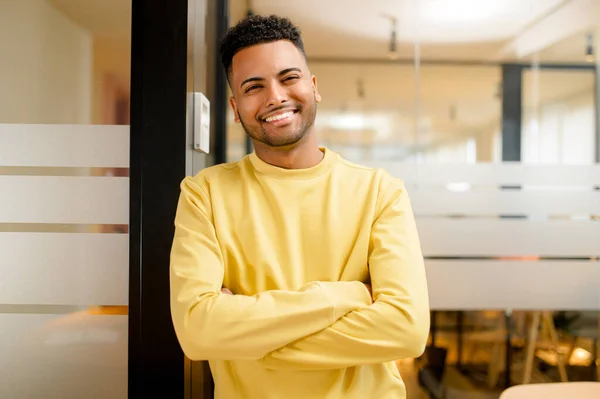 Smiling indian businessman standing in modern open space office and looking at the camera. Young positive male student, proud and successful multiracial small business owner
