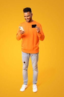 Neşeli yakışıklı adam cep telefonuyla uzun zamandır beklenen alışverişi yapıyor, elinde kredi kartı var ve ekrana bakıyor. Orta Doğulu mutlu adam internetten yemek sipariş ediyor.