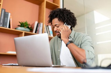 Yakışıklı kıvırcık saçlı Afro-Amerikalı ofis masasında dizüstü bilgisayarın önünde oturuyor ve telefon görüşmesi yapıyor. İş adamı, ortakları ya da müşterileriyle akıllı telefondan konuşuyor.
