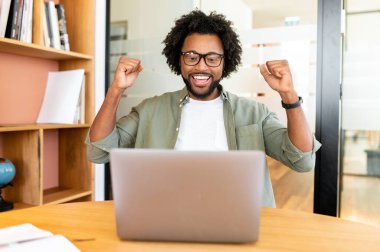 Mutlu Afro-Amerikalı adam zaferi kutluyor laptopun önünde birlikte çalışıyoruz, şanslı işadamı bağırıyor evet, iyi haberler, iş teklifi