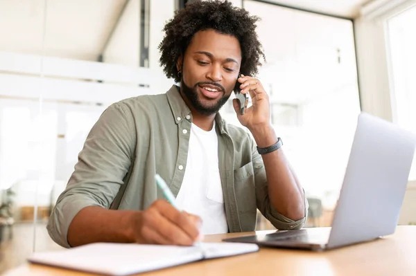 アフリカ系アメリカ人のビジネスマンが電話でオフィスに座ってメモを書き フレンドリーな男性従業員が顧客と携帯電話で話し ノートパソコンの前でスマートフォンでチャットします — ストック写真