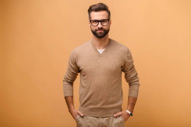 Bej renkli kazak giyen yakışıklı sakallı adam ceplerinde el ele tutuşuyor, şık gözlüklü çekici adam kameraya bakıyor hoş bir gülümsemeyle, izole edilmiş stüdyo çekimiyle.