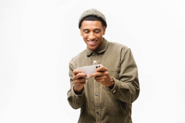 Yakışıklı, kıvırcık, Afro-Amerikan, genç hippi, yatay pozisyonda duruyor. Akıllı telefonu heyecanlı bir suratla kullanıyor, video oyunu oynuyor, video izliyor. Beyaz arkaplanda stüdyo görüntüsü