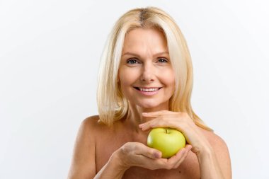 Yüzünün yanında yeşil elma tutan, sağlıklı ve güzel orta yaşlı bir kadın, kameraya bakıyor ve gülümsüyor, stüdyo güzelliği portresi beyaza izole edilmiş.