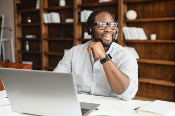 Serious Positive and cheerful african-american businessman in headset sitting in front of the laptop and talking online. Happy male employee looking away, having pleasant conversation