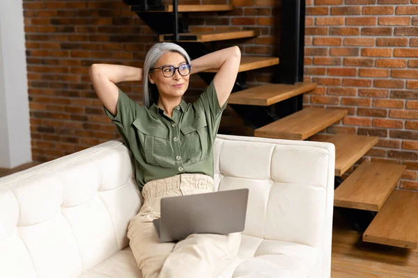 现代60多岁的女性在家里享受周末的闲暇 在电脑上浏览无线互联网 快乐的老妇人坐在客厅的沙发上 面带微笑地看着远方 — 图库照片