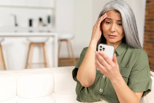 灰白头发的垂头丧气的成熟女人坐在沙发上 拿着智能手机 老年女性没有足够的钱进行网上交易 财务问题 — 图库照片