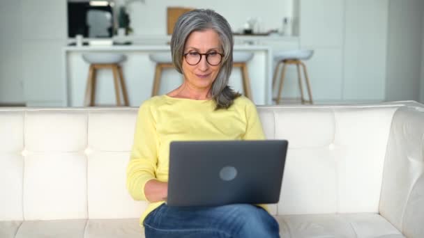 漂亮的中年妇女坐在白色的沙发上 用笔记本电脑 开心地笑着 在电脑上打字 发短信 阅读新闻 找工作 远程办公 — 图库视频影像