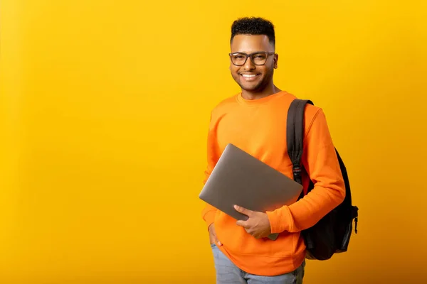 Happy excited foreign male student with backpack standing isolated on yellow and holding laptop, arab guy in glasses ready to studying, back to school concept