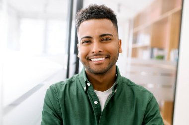 Arkadaşça geniş dişli gülüşü olan mutlu Hintli genç bir adamın yakın plan portresi, gündelik yeşil tişörtlü Latin bir adam kameraya bakar, çalışan profili fotoğrafı