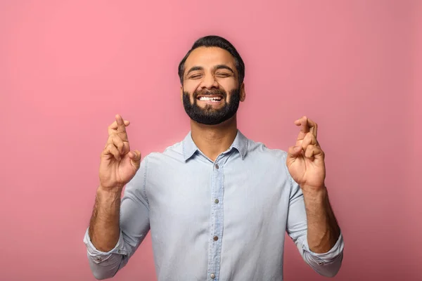 Wish me luck. Smiling young Indian bearded man holding fingers crossed, eyes closed in hope, multiracial guy really wants to be lucky, isolated on pink wall