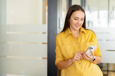 Ofiste akıllı telefonu tutan ve koridorda dinlenirken mesaj yazan olumlu bir kadın. İş adamları konsepti