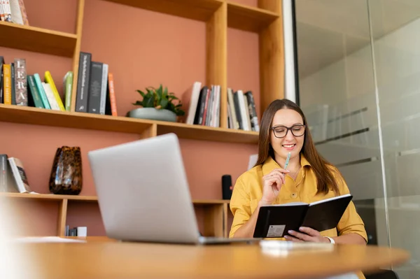 魅力的な女性は ノートパソコンのテーブルに座ってノートを取り 電子学習上のオンラインウェビナーを見て 女性研究室での仕事の分析 — ストック写真
