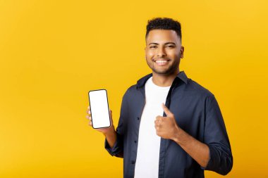 Kameraya bakan mutlu Hintli genç adam, elinde telefon ekranı boş olan akıllı telefonu tutuyor ve yeni tanıtım işareti olarak baş parmağını gösteriyor, sarı ekranda izole edilmiş yeni bir uygulama öneriyor.