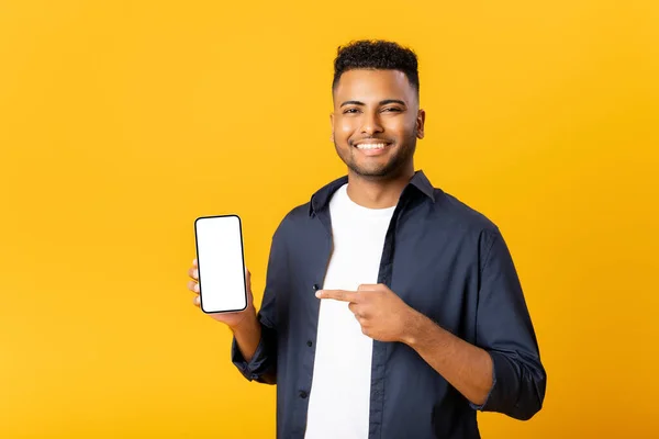 Happy excited young indian man looking at camera pointing finger at empty phone screen, paying attention at copy space, advertising new promotion, presenting sale offer standing isolated on yellow