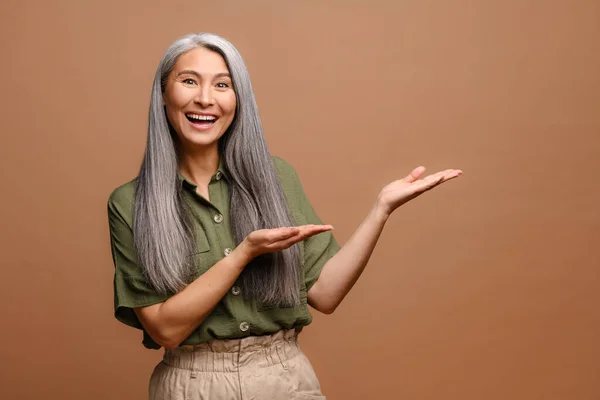 興奮した灰色の髪50アジアの女性ポインティング距離で両方の手上のブランクコピースペース ショー場所のためにあなたの広告隔離された上茶色の背景 — ストック写真