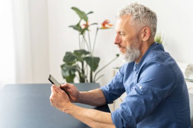 Ofisteki masada oturan beyaz saçlı, sakallı yaşlı adam akıllı telefon kullanıyor. İnternette haber okuyan, telefonda mesajlaşan akıllı ve rahat gömlekli olgun bir erkek. Yan görünüm