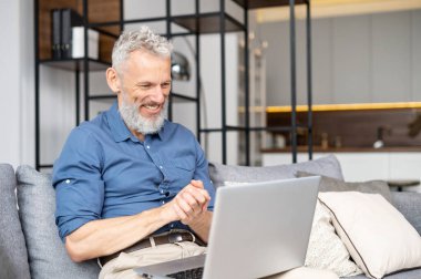 Modern orta yaşlı sakallı adam evdeki koltukta dizüstü bilgisayar kullanıyor boş zamanlarını internette film izleyerek geçiriyor. Olgun erkek evden uzakta çalışıyor, ekrana bakıyor ve gülümsüyor.