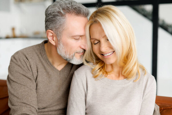 Cheerful and happy middle-aged spouses enjoying time together. Handsome grey-haired man hugging gently beautiful good-looking woman, their eyes are closed. Love and bond concept. Close-up portrait