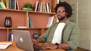 Uzaktan temastayız. Kendine güvenen Afro-Amerikan erkek çalışanı iş arkadaşlarıyla sanal iletişim kurmak için online platform kullanıyor, webcam 'e el sallıyor, video görüşmesi yapıyor, arka planda kitaplıklar yapıyor.,