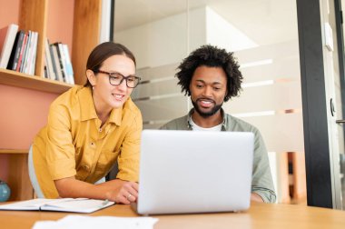 İki mutlu iş arkadaşı Afrika kökenli Amerikalı erkek ve beyaz kadın dizüstü bilgisayara bakıyor ve ofis lobisinde oturuyor.